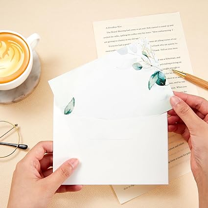 Persona escribiendo unas tarjetas para felicitación o celebración manuscritas con sobre decorado con hojas verdes, sobre una mesa con café y gafas, transmitiendo elegancia y calidez.
